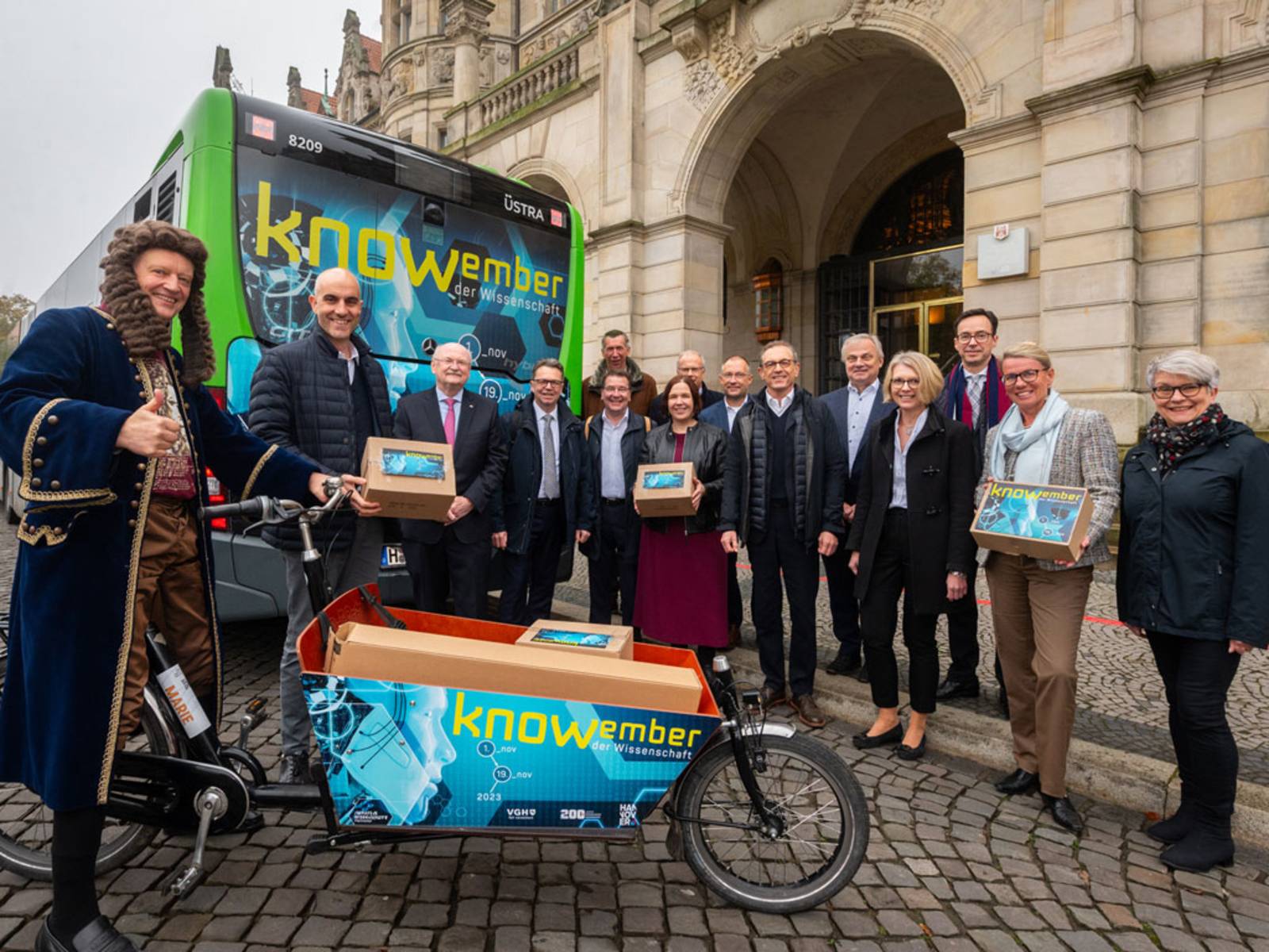 Menschengruppe mit Bus und Lastenrad. Auf dem Bus und dem Lastenrad steht "knowember".