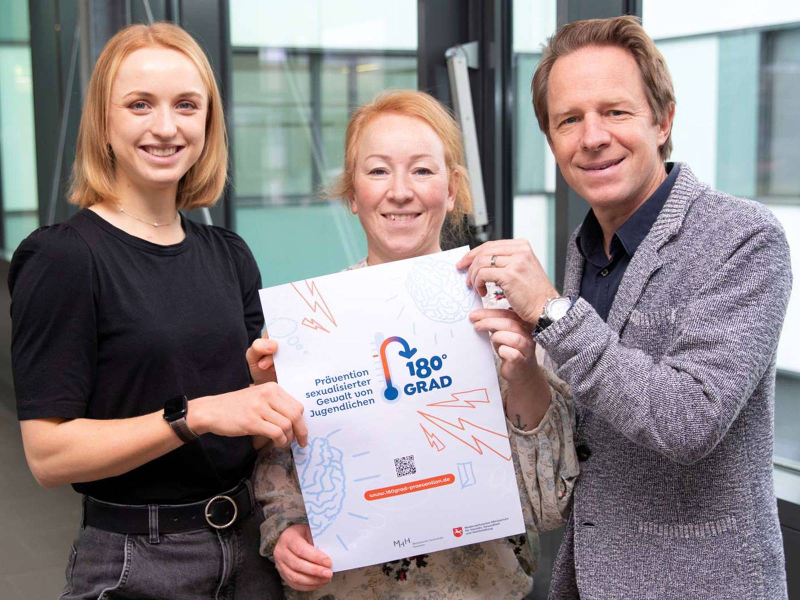 Zwei Frauen und ein Mann stehen mit einem Plakat in der Hand in einem Klinikgang.