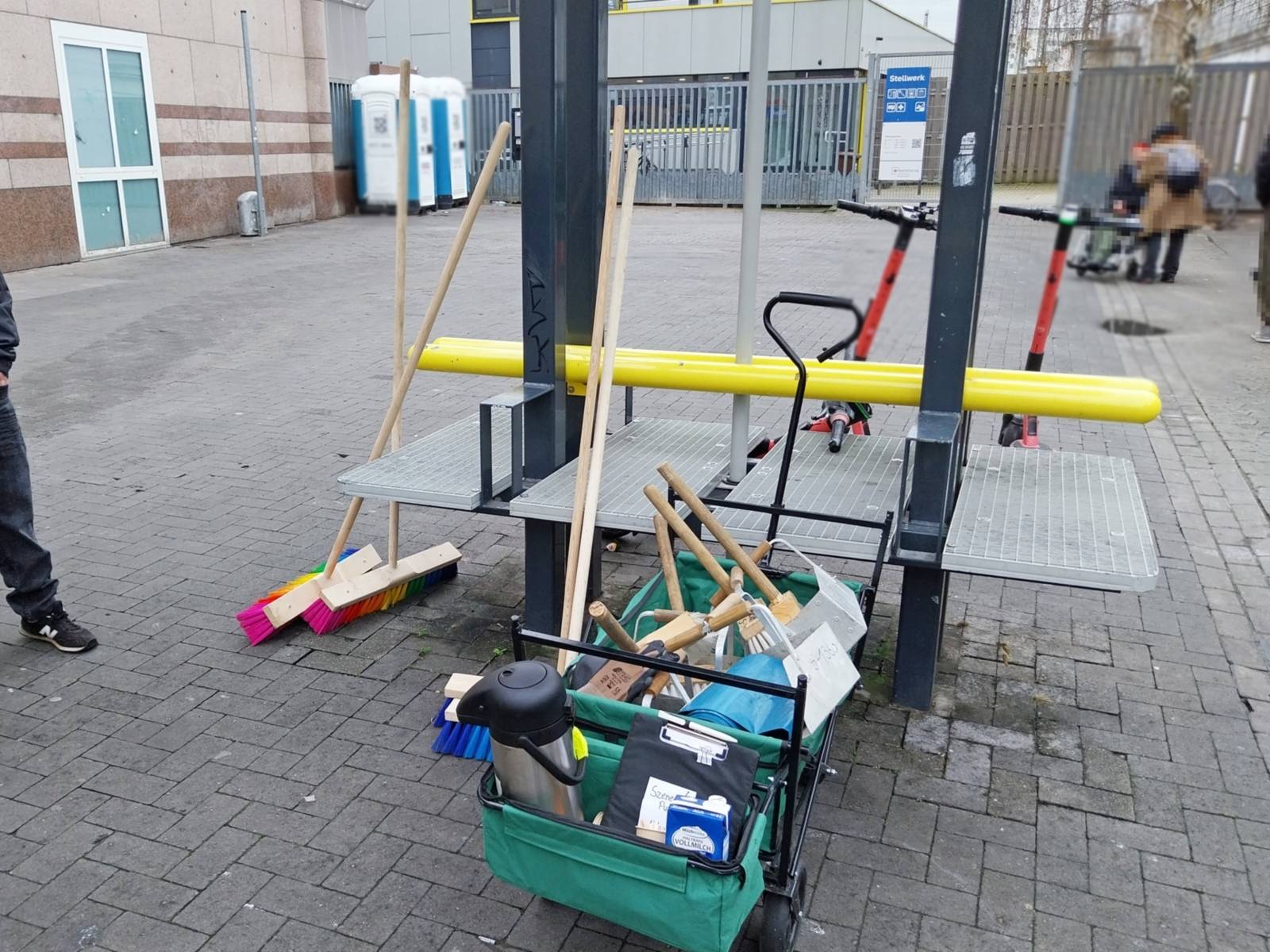 Eine Transportkarre mit Putzzeug udn einer Kaffeekanne steht draußen auf Pflastersteinen, links stehen  zwei Besen mit regenbogenfarbenen Borsten.