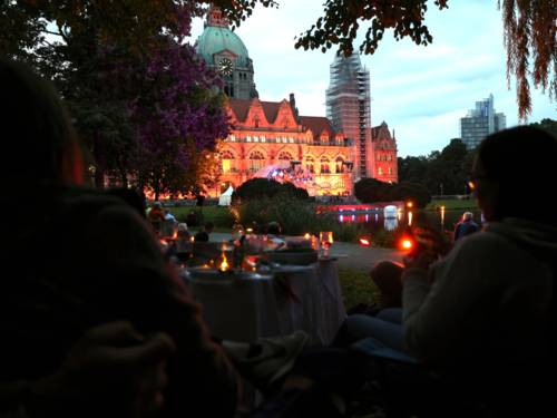 Menschen, die abends in einem Park sitzen. Sie blicken auf eine beleuchtete Bühne vor dem Neuen Rathaus.