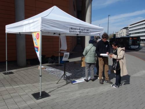 Es sind vier Personen auf dem Bild zu erkennen, die sich jeweils zu zweit unterhalten. Sie stehen unter, beziehungsweise bei einem Pavillon mit der Aufschrift "Masterplan für das nördliche Bahnhofsviertel".