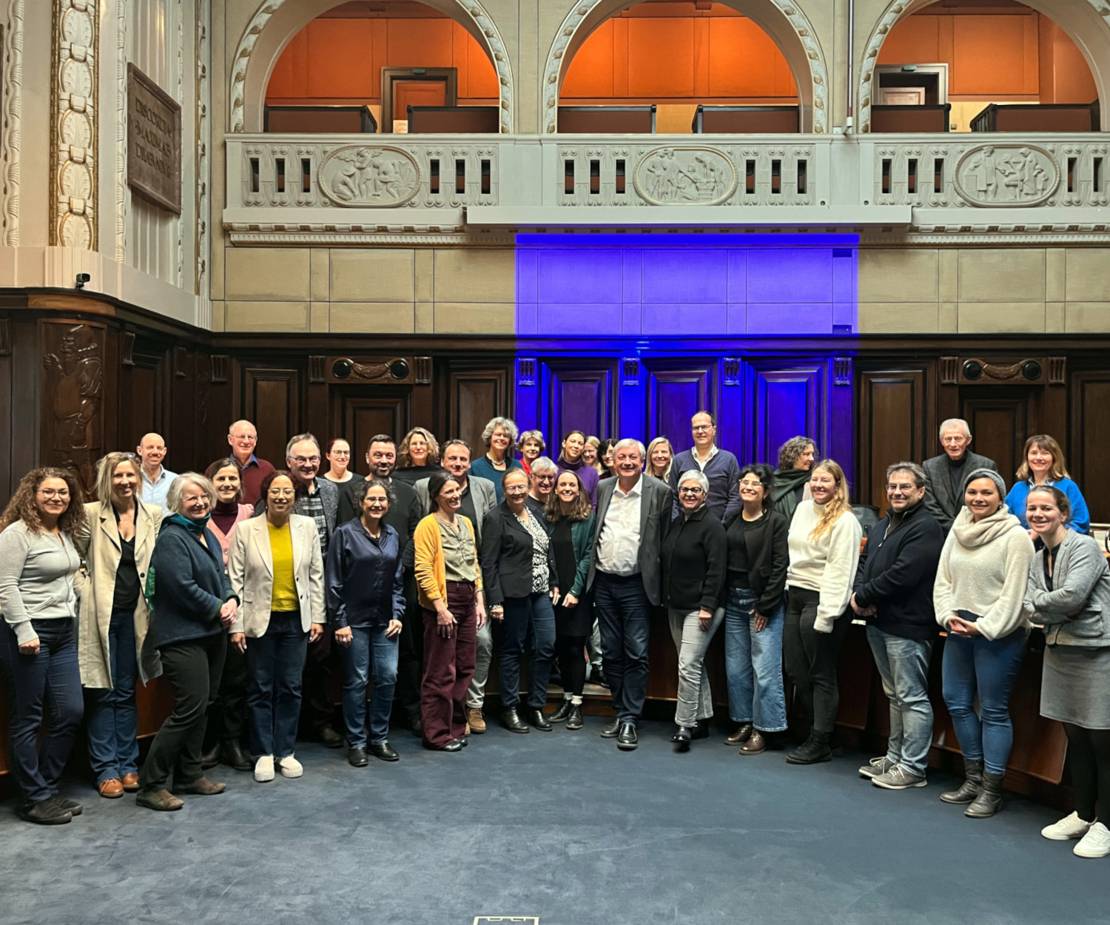 34 Personen posieren in einem angedeuteten Halbkreis für ein Gruppenfoto. Im Hintergrund die dunkle Vertäfelung und der helle Zuschauer und Zuschauerinnen Balkon des Hodlersaals im Neuen Rathaus.