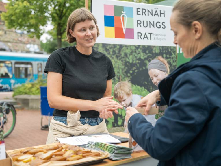 Eine Frau hinter einem Tisch redet mit einer anderen Frau ihr gegenüber. Hinter der ersten steht ein Schild mit der Aufschrift "Ernährungsrat Hannover".