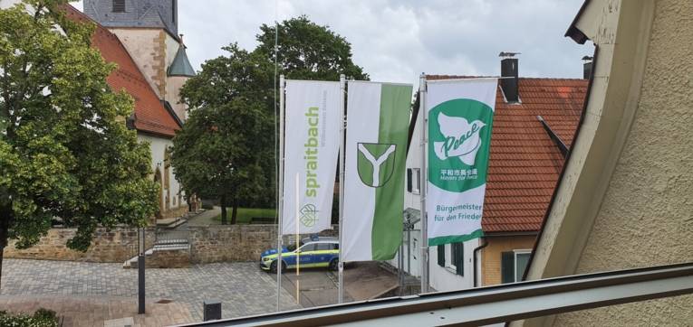 Die Friedensflagge der Mayors for Peace ergänzt das Gemeindewappen und Logo von Spraitbach